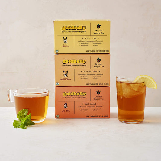 Goldholly three-roast bundle pack featuring stacked boxes of Ocelot Light, Panther Medium, and Red Wolf Dark Roast with glass mugs of amber yaupon tea. Complete collection showcases the full spectrum of America's native caffeine flavors from bright to bold.