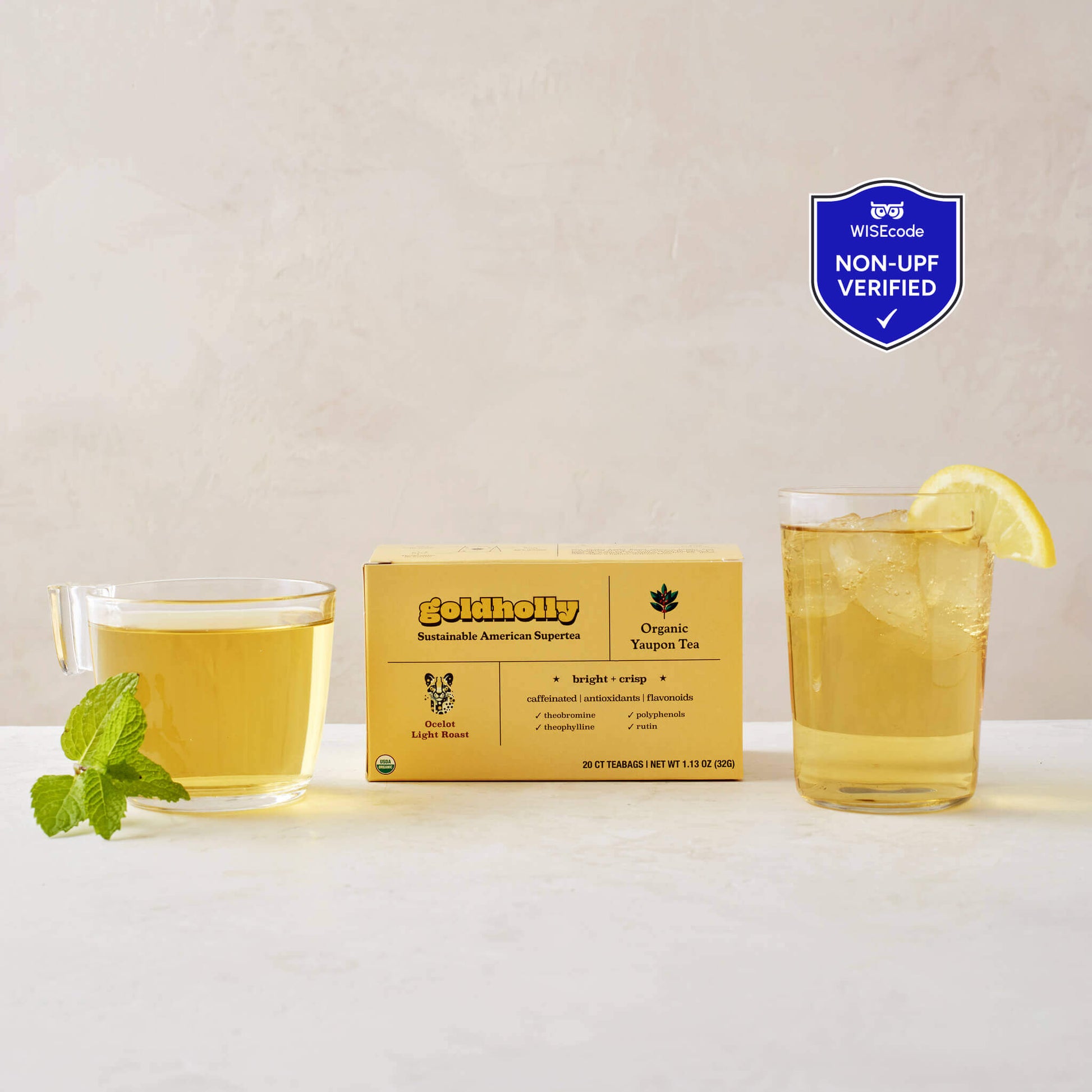 Goldholly Ocelot Light Roast box with clear glass mugs filled with golden yaupon tea on clean white marble surface. Fresh mint garnish and lemon slice enhance the bright, crisp flavor profile of America's only native caffeinated plant, with the WISEcode Non-UPF verified shield placed in the upper right corner of the image.