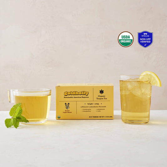 Goldholly Ocelot Light Roast box with clear glass mugs filled with golden yaupon tea on clean white marble surface. Fresh mint garnish and lemon slice enhance the bright, crisp flavor profile of America's only native caffeinated plant, with the USDA Certified Organic seal and WISEcode Non-UPF verified shield in the upper right side of the image.