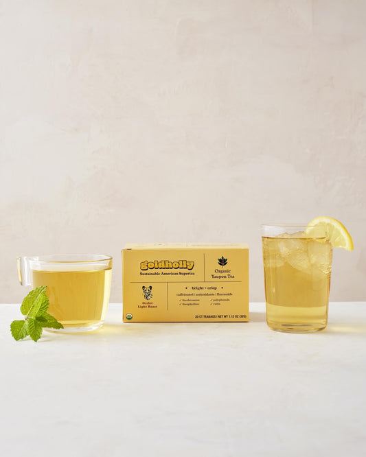 Elegantly styled Goldholly Ocelot Light Roast box with clear glass mugs filled with golden yaupon tea on clean white marble surface. Fresh mint garnish and lemon slice enhance the bright, crisp flavor profile of America's only native caffeinated plant.