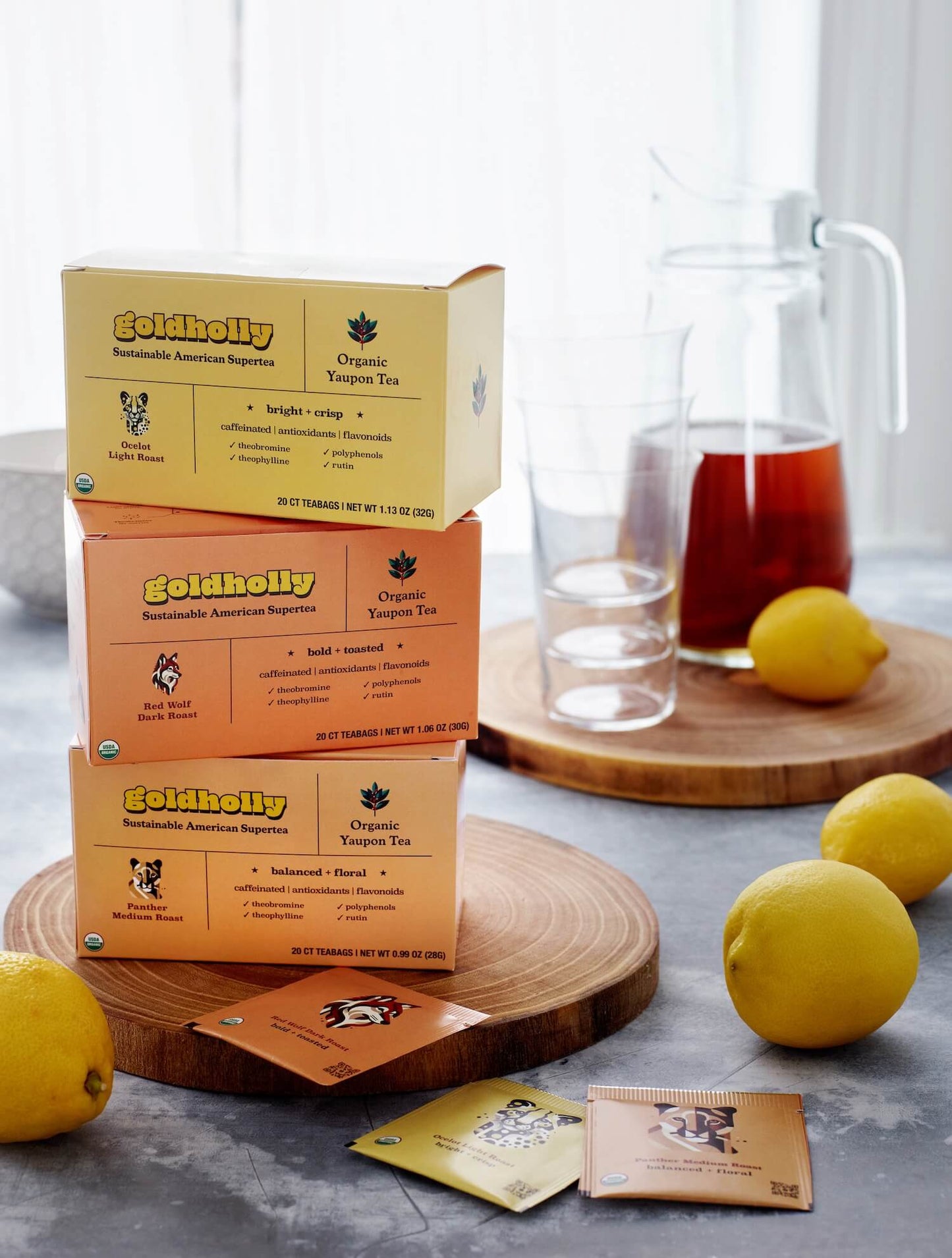 Bright kitchen scene with Goldholly three-roast bundle pack stacked on wood cutting board beside glass pitcher of iced tea and fresh lemons. Natural morning light illuminates the complete collection of sustainable American yaupon varieties.
