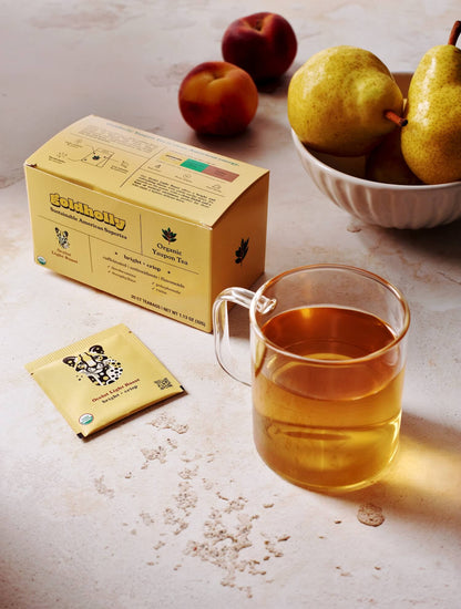Goldholly Ocelot Light Roast box with steaming glass mug of amber yaupon tea surrounded by fresh peaches and pears. Scattered loose tea leaves reveal the natural, earthy character of this sustainable American supertea.