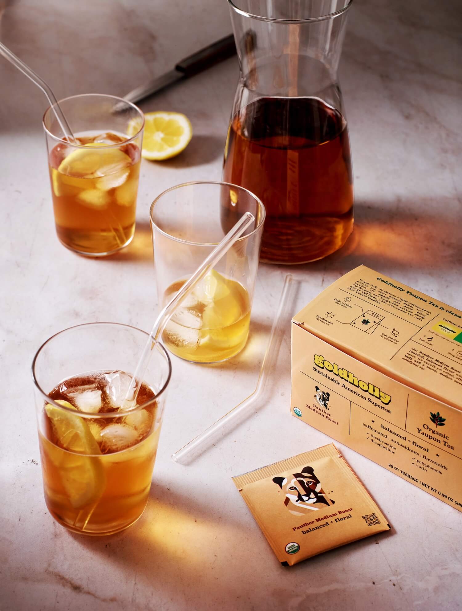 Sophisticated iced tea service featuring Goldholly Panther Medium Roast with glass pitcher, tumbler glasses, and golden honey straws. Crystal-clear glassware highlights the rich amber color of cold-brewed yaupon.