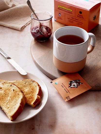Cozy breakfast setting with Goldholly Red Wolf Dark Roast tea in two-toned ceramic mug paired with jam and toast. Warm morning light creates perfect atmosphere for bold, toasted yaupon flavors.