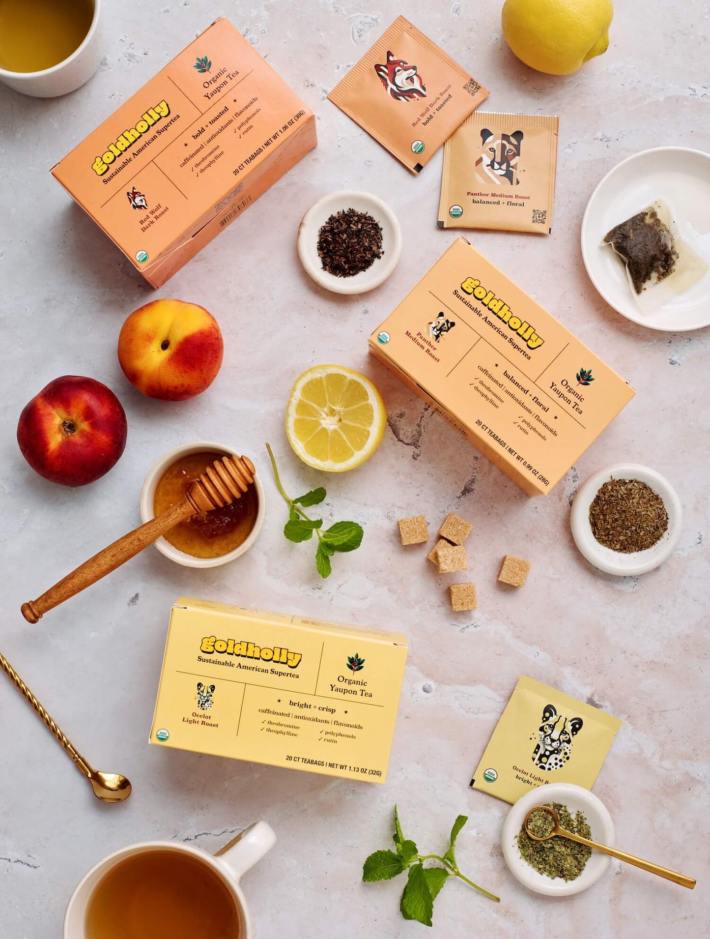 Goldholly three-roast bundle pack varieties displayed with loose tea leaves, honey dipper, fresh fruit, and raw sugar cubes on marble surface. Complete flavor journey from bright Ocelot to bold Red Wolf showcases yaupon's versatile taste profiles.