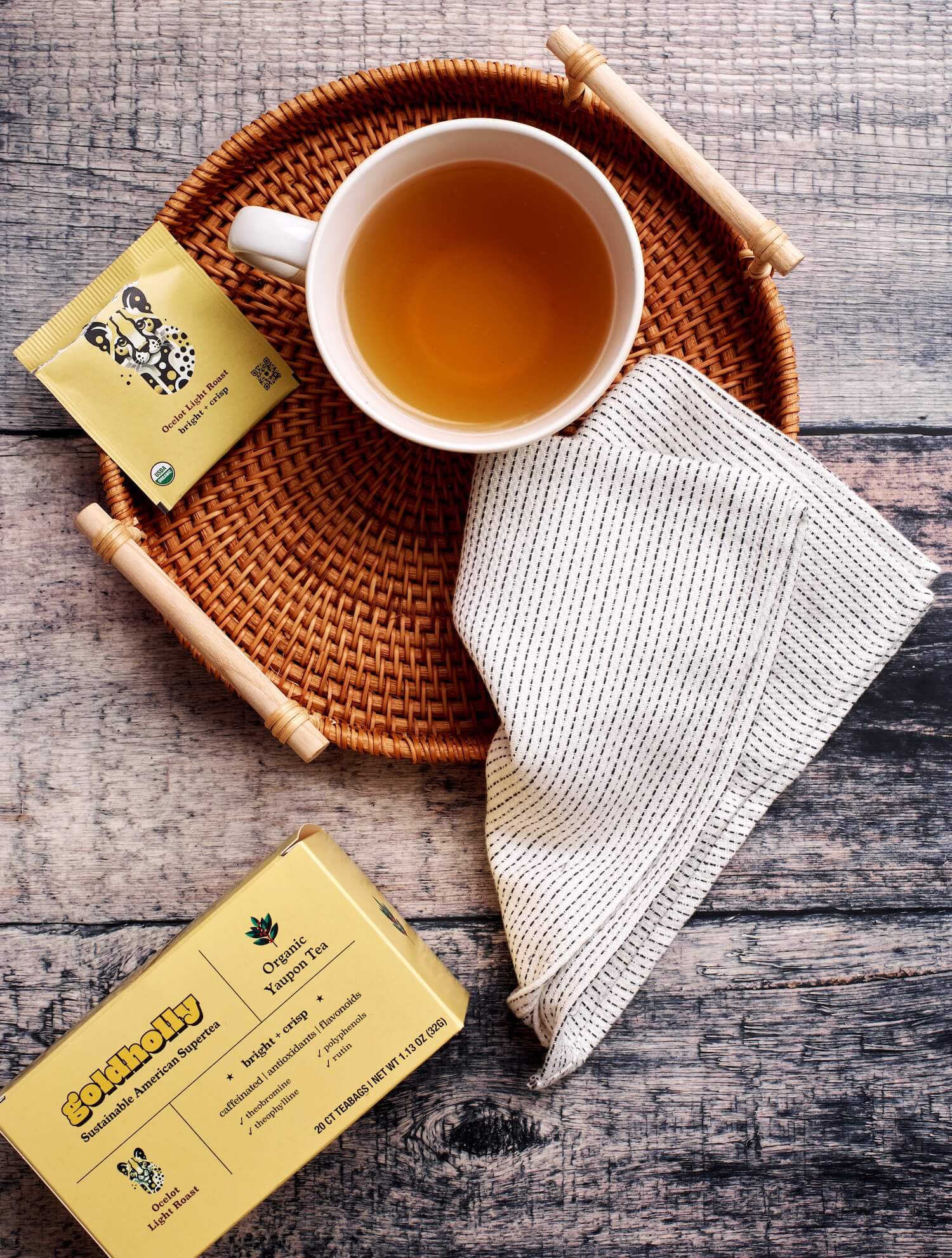 Cozy wicker basket holds steaming cup of golden Goldholly yaupon tea with natural linen cloth and teabag packet. Rustic wood surface creates warm, inviting atmosphere perfect for savoring smooth, native caffeine.
