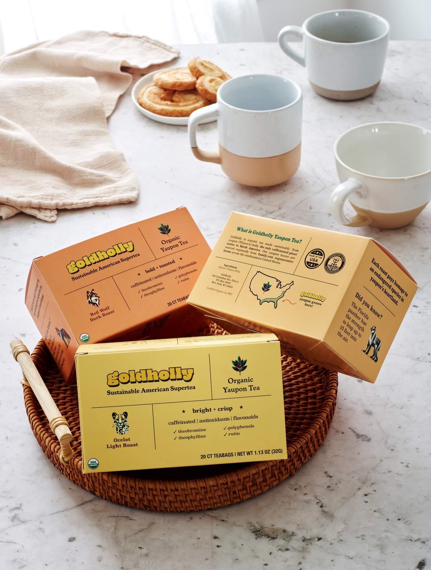 Goldholly three-roast bundle pack beautifully arranged in wicker basket with ceramic mugs and linen cloth. Perfect gift presentation featuring America's complete collection of organic, sustainably-sourced yaupon tea varieties.