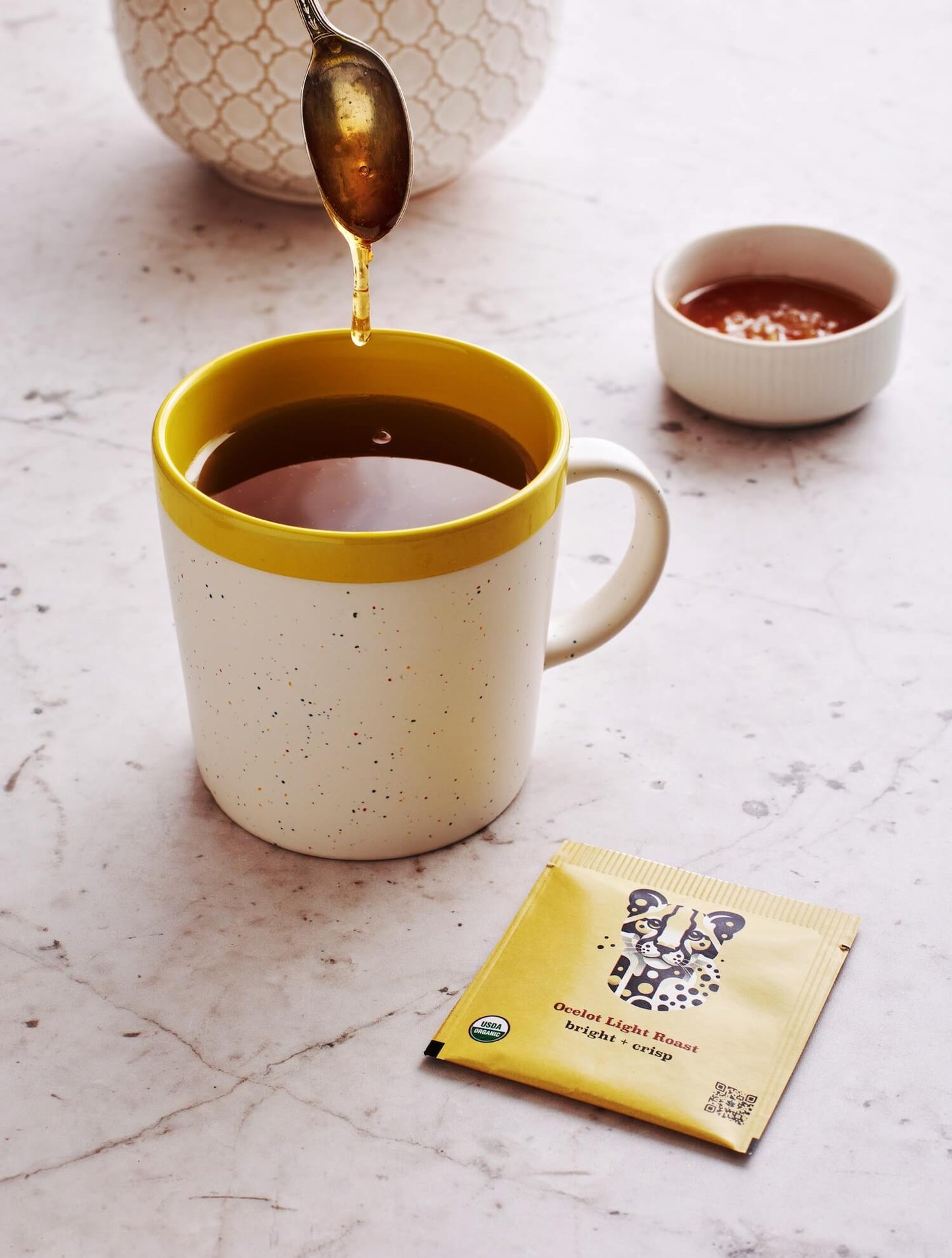 Golden honey drizzles into speckled ceramic mug of rich Goldholly yaupon tea. Yellow-rimmed cup showcases the amber brew while Ocelot Light Roast packet promises bright, naturally sweet flavor notes.