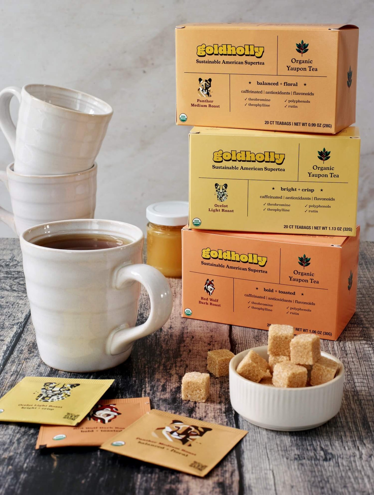 Goldholly bundle pack tower on rustic wood surface with steaming white ceramic mug and raw sugar cubes. Morning ritual setup showcases the complete range of native American caffeine from light to dark roast.