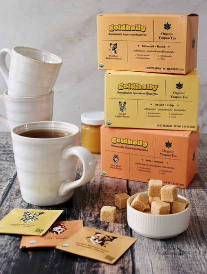 Goldholly bundle pack tower on rustic wood surface with steaming white ceramic mug and raw sugar cubes. Morning ritual setup showcases the complete range of native American caffeine from light to dark roast.