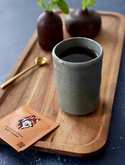 Goldholly Red Wolf Dark Roast teabag beside modern speckled ceramic cup with yaupon tea on warm wood serving board. Contemporary styling highlights the bold, toasted character of America's native caffeine.