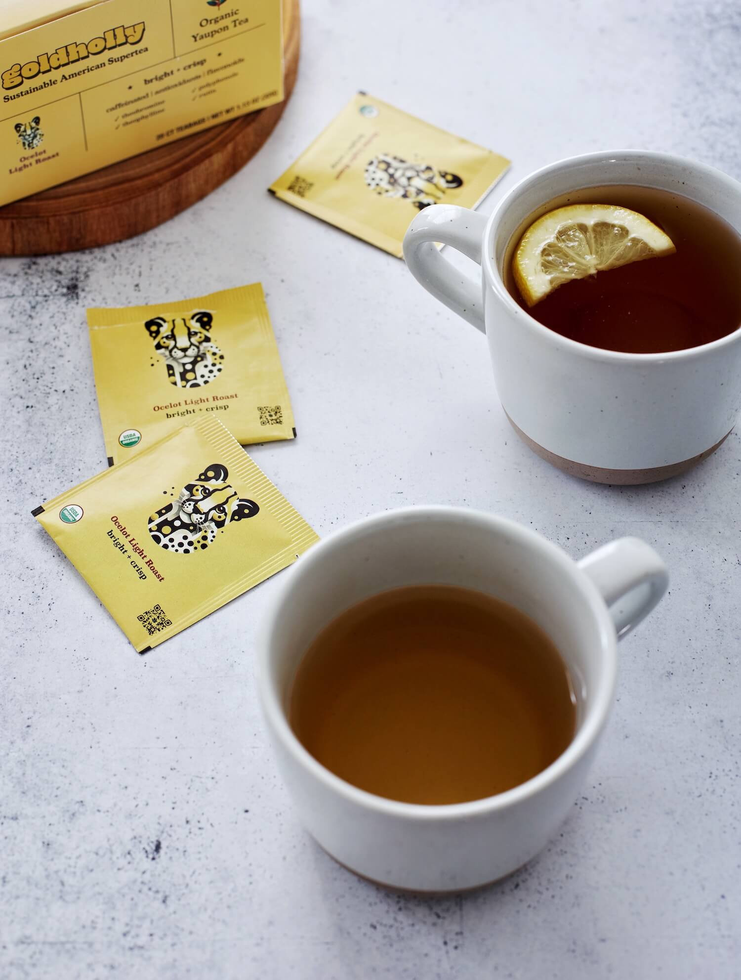 Two white mugs filled with golden Goldholly Ocelot Light Roast yaupon tea on speckled marble surface. Bright yellow teabag packets and wooden cutting board create inviting morning ritual with America's sustainable caffeine.