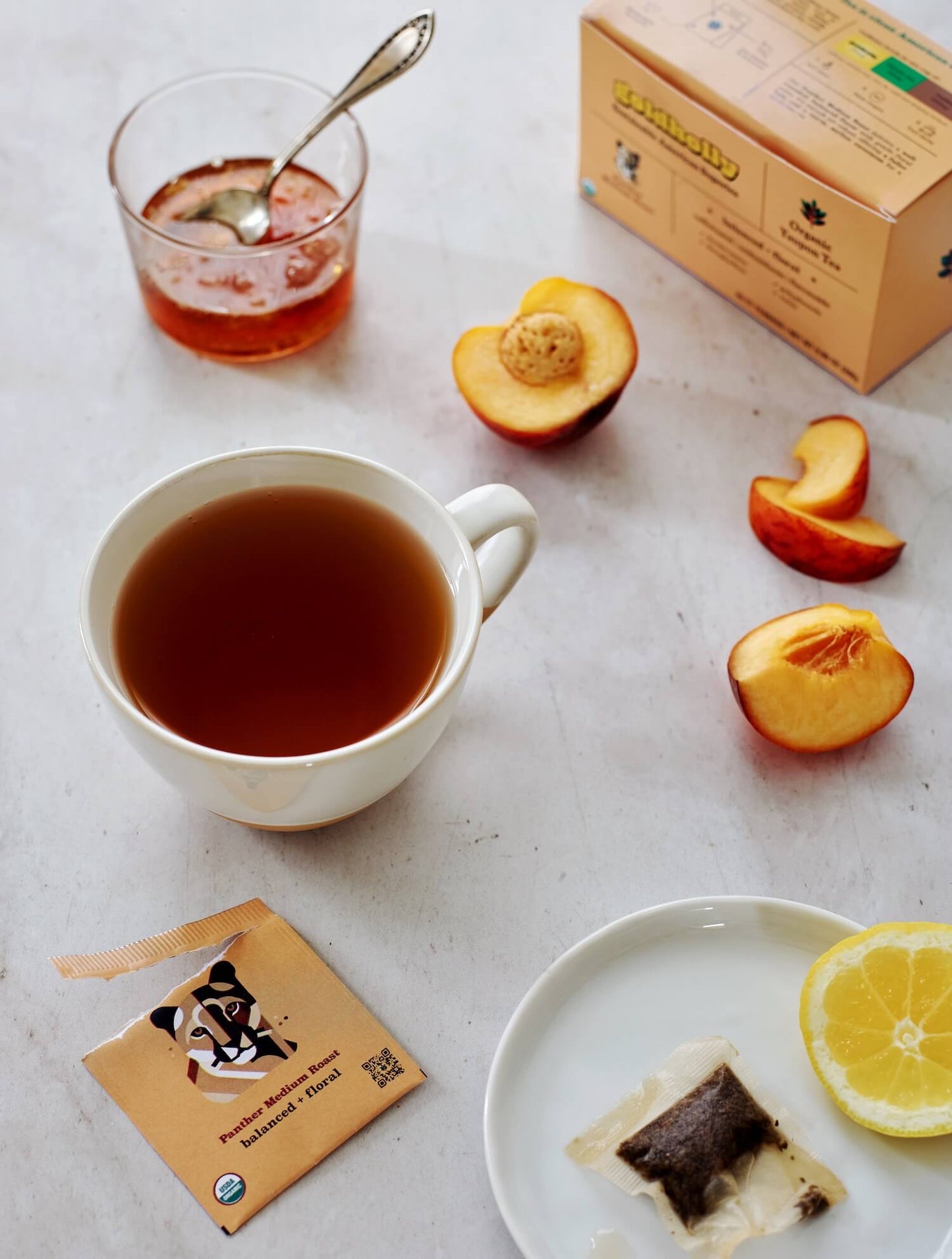 Goldholly Panther Medium Roast cup surrounded by fresh peach slices and teabag on white marble. Natural sweetness of summer fruit complements the balanced, floral notes of organic yaupon tea.