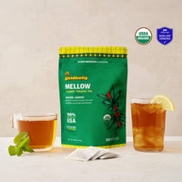 Goldholly Mellow organic yaupon tea pouch in vibrant green with Malted + Earthy flavor tag, flanked by a warm cup of brewed tea with mint and an iced glass with lemon, with three teabags. USDA Organic certified and WISEcode Non-UPF verified.--1
