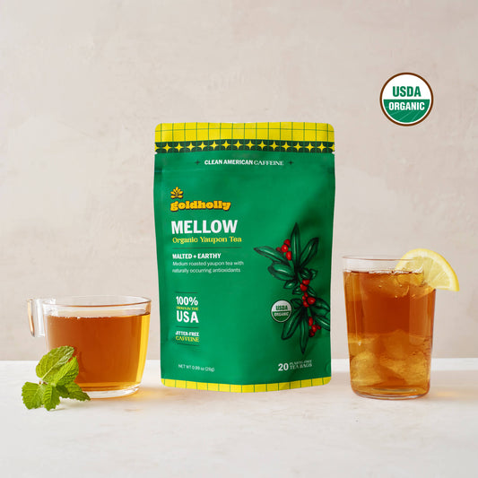 Goldholly Mellow organic yaupon tea pouch in vibrant green with Malted + Earthy flavor tag, flanked by a warm cup of brewed tea with mint and an iced glass with lemon, USDA Organic certified
