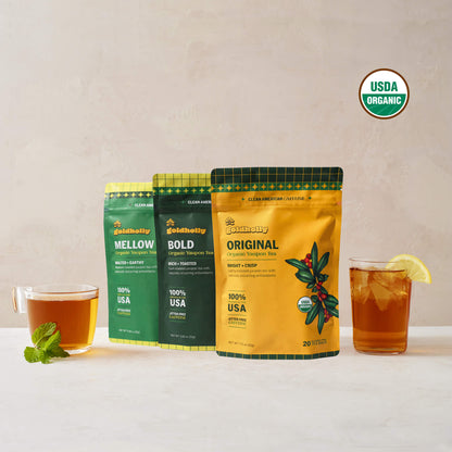 Goldholly variety bundle with Original, Bold, and Mellow organic yaupon tea pouches flanked by a warm cup of brewed tea and an iced glass with lemon, USDA Organic certified
