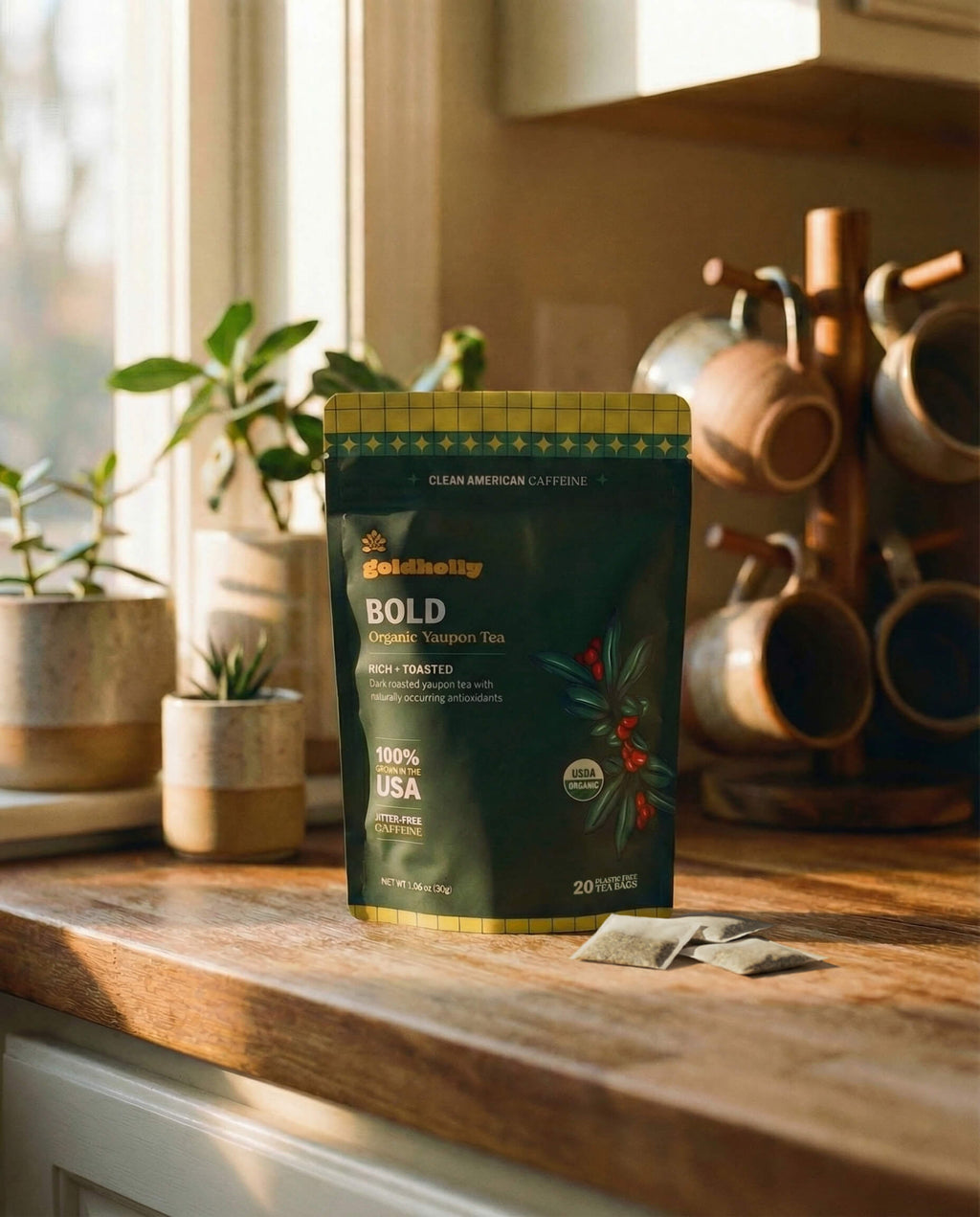 Goldholly Bold organic yaupon tea pouch on a warm sunlit kitchen counter with loose teabags beside it, copper mug tree and green plants in the background--6