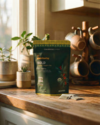 Goldholly Bold organic yaupon tea pouch on a warm sunlit kitchen counter with loose teabags beside it, copper mug tree and green plants in the background--6
