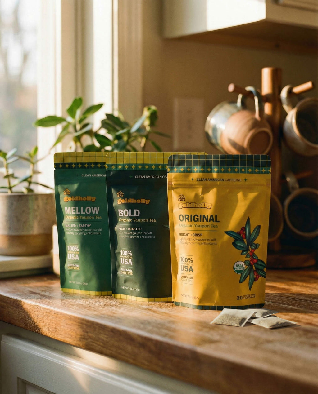 Three Goldholly organic yaupon tea pouches, Original, Bold, and Mellow, lined up on a warm sunlit kitchen counter with loose teabags, copper mugs, and green plants--6
