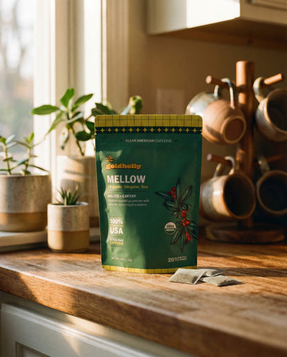 Goldholly Mellow organic yaupon tea pouch on a warm sunlit kitchen counter with loose teabags beside it, copper mug tree and green plants in the background