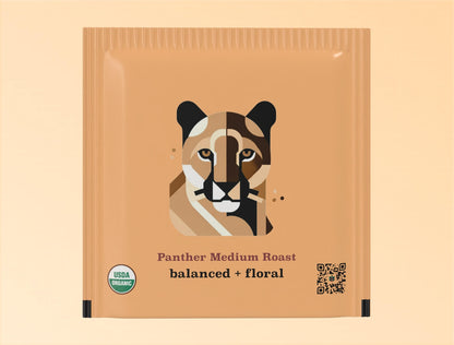 Individual teabag packaging for Goldholly's Panther Medium Roast yaupon tea. The tan-colored sachet features a geometric, color-blocked illustration of a panther face with distinctive markings in shades of brown, black, and cream. Below the illustration are the words 'Panther Medium Roast' and 'balanced + floral' in brown typography. The package includes a USDA Organic certification seal and QR code.