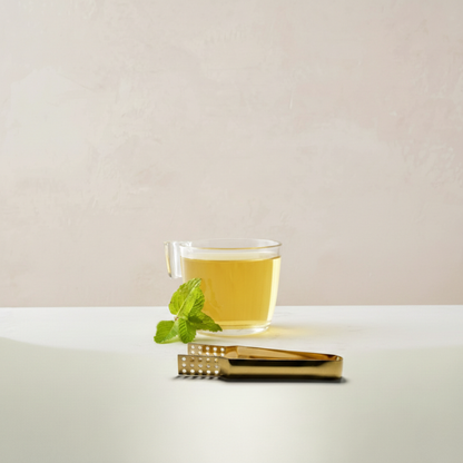 Teabag tongs next to a clear glass of Goldholly Yaupon, with a sprig of mint alongside.