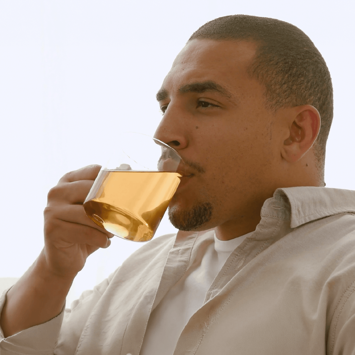 A man in a light beige button-up shirt drinking from a clear glass mug containing amber-colored yaupon tea, set against a bright, overexposed white background.