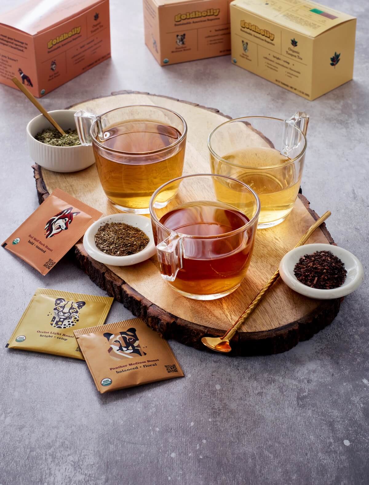 Artisanal tea tasting display featuring three glass mugs of Goldholly yaupon tea in varying amber hues on rustic live-edge wood board. Loose tea leaves in white ceramic bowls showcase the texture differences between bright Ocelot Light Roast, balanced Panther Medium Roast, and bold Red Wolf Dark Roast varieties. Golden honey spoon and organic teabag packets complete this inviting exploration of America's native caffeine alternatives.