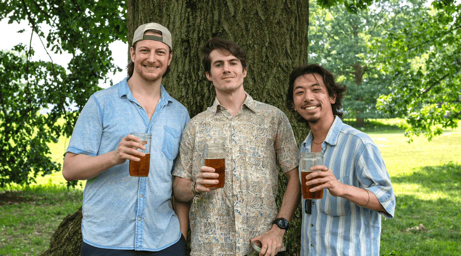 Three men standing outdoors in front of a large tree, each holding a glass of iced yaupon tea. They are wearing casual short-sleeve button-up shirts in light blue, beige, and blue-and-white stripes.